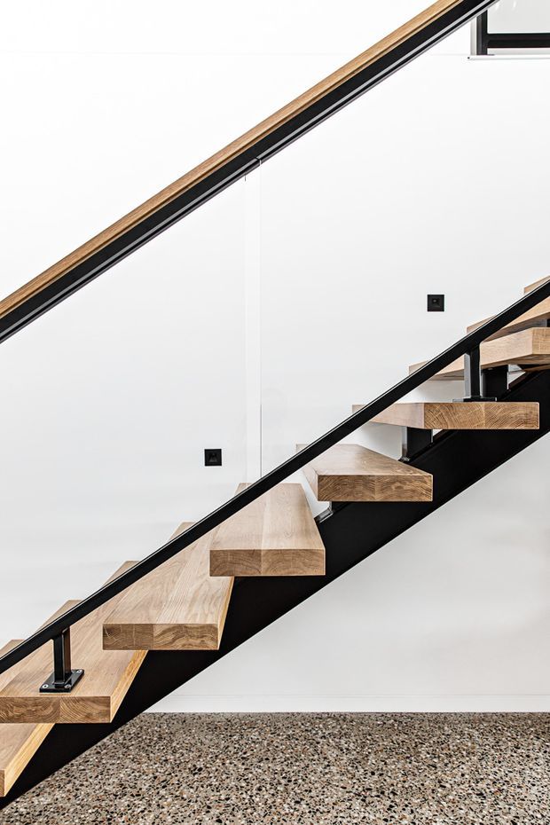A close-up architectural photograph of a contemporary open-riser staircase. Thick natural timber treads are cantilevered from a black steel structural spine, with slim black metal handrails and posts. The staircase is set against smooth white walls and a terrazzo floor, creating a clean, minimalist interior composition.