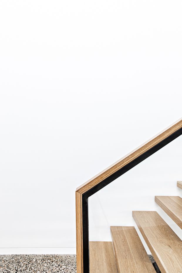 Minimal architectural detail showing a floating timber staircase with light wood treads, a black-framed glass balustrade, white walls, and a terrazzo-style floor. The composition emphasizes clean lines and the junction between the stair and wall.