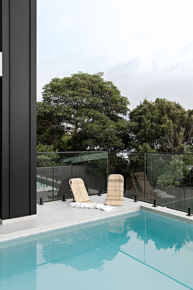A contemporary outdoor pool area viewed from the water, with a turquoise swimming pool, pale tiled pool surround, frameless glass balustrade, and two woven sun loungers. Dense mature trees form a leafy backdrop, while a dark vertical building facade is visible at the left.
