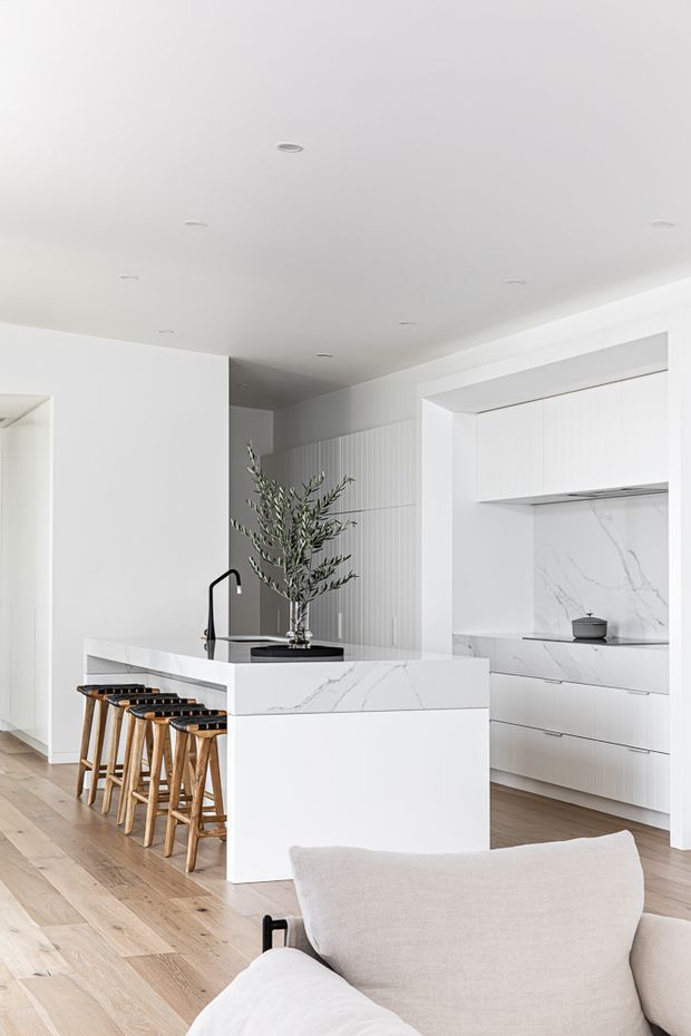 Bright, refined contemporary kitchen with a large white island, integrated cabinetry, veined stone surfaces, a black gooseneck tap, timber bar stools, and pale timber flooring. A partial upholstered lounge chair is visible in the foreground, while vertical wall panelling and recessed ceiling lights add subtle detail.