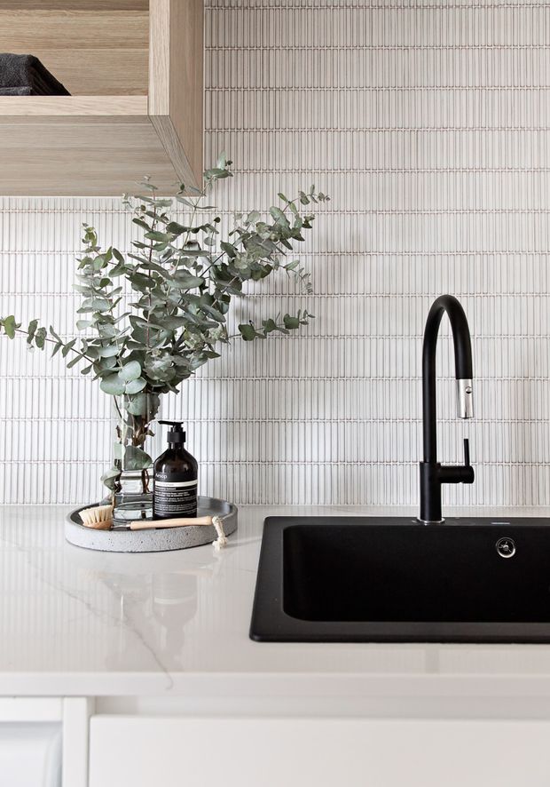Close-up of a refined contemporary kitchen sink area. A matte black undermount sink and tall black pull-out tap sit within a pale stone benchtop, backed by a vertically ribbed light tile splashback. Light timber cabinetry and open shelving appear above, with a vase of eucalyptus and black soap dispenser adding natural and dark accents.