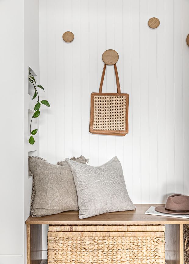 A bright, styled built-in nook featuring a timber bench seat with neutral textured cushions, woven basket storage below, and a cane-front hanging bag mounted against a white vertical-panel wall. A trailing plant adds a natural accent, with a hat and magazine resting on the bench.