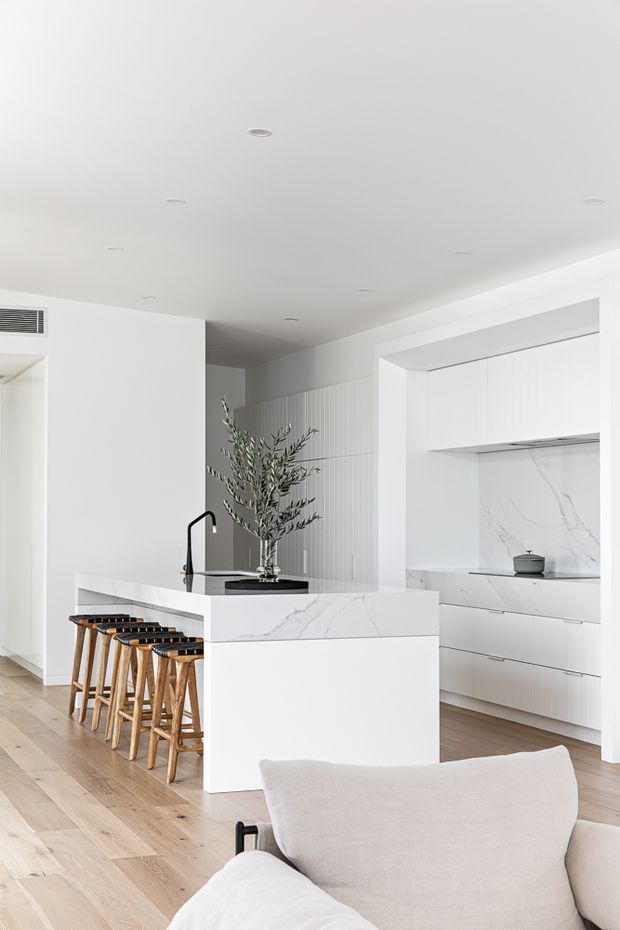Bright, minimalist contemporary kitchen viewed from an adjoining living area. The room features a large white stone island with integrated sink and black tap, timber bar stools, white vertically fluted cabinetry, a marble-look splashback and benchtops, pale timber flooring, and recessed ceiling lights. A light upholstered sofa is visible in the foreground.