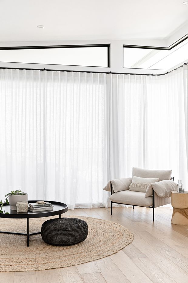 Bright, serene living room with floor-to-ceiling sheer white curtains, pale timber flooring, a low black round coffee table, a woven round rug and pouf, and a neutral upholstered armchair. High-level clerestory-style glazing and recessed ceiling lights enhance the airy minimalist setting.