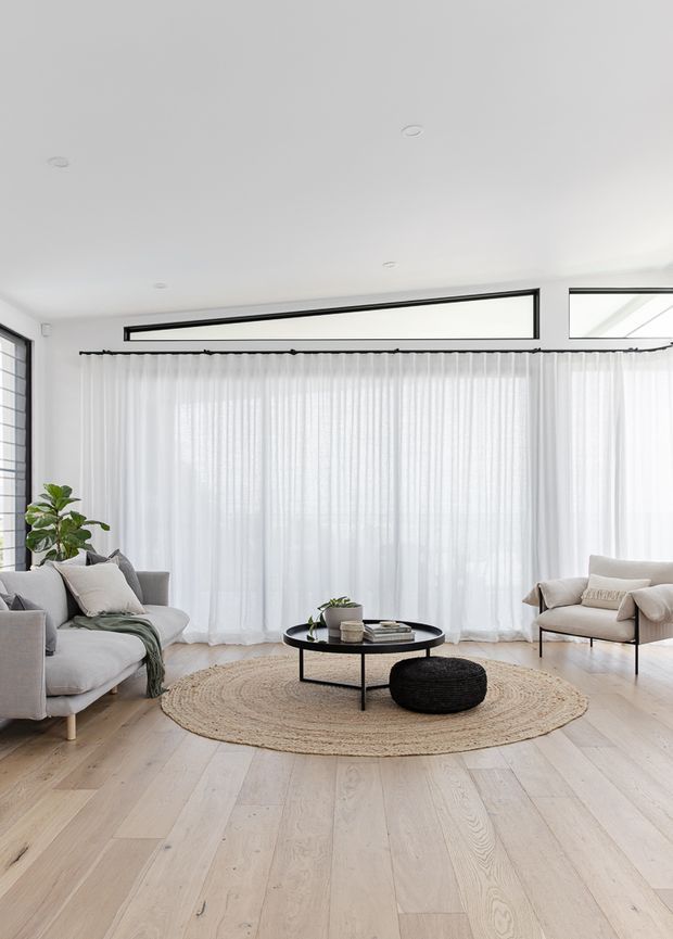 Bright, airy contemporary living room with pale timber flooring, floor-to-ceiling sheer white curtains, expansive black-framed clerestory-style glazing, two light upholstered sofas, a round black coffee table, woven round rug, and black ottoman. The room has a calm, minimalist neutral palette with abundant natural light.