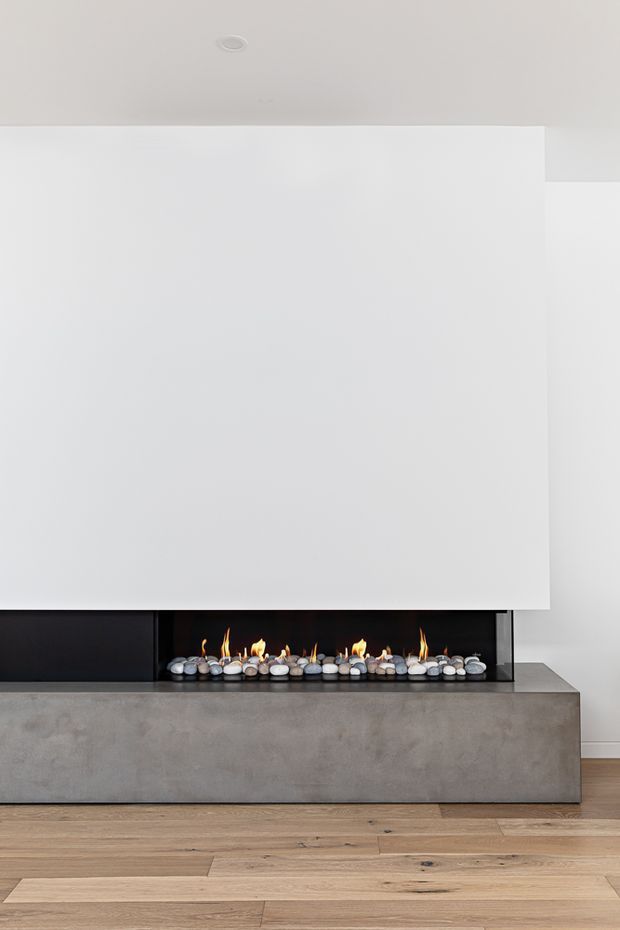Minimalist interior detail featuring a long linear fireplace set into a dark surround, with a substantial smooth grey concrete hearth or plinth. The fireplace is decorated with rounded stones and visible flames. A large plain white wall and ceiling dominate the composition, with warm natural timber flooring below.