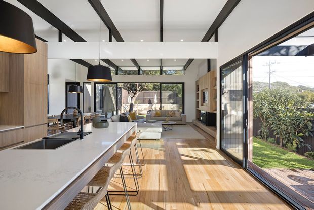A bright, open-plan contemporary kitchen and living area with a long white stone island, integrated sink, black tapware, and woven bar stools. Warm timber cabinetry and timber flooring contrast with exposed dark ceiling beams, black pendant lights, and extensive floor-to-ceiling glazing opening to a landscaped garden and deck. A lounge area and fireplace are visible beyond the kitchen, with strong natural light creating an airy indoor-outdoor connection.