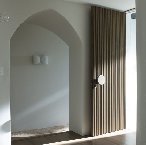 Minimalist interior entry or hallway with a sculptural arched opening, pale plaster walls, herringbone timber flooring, and a tall dark timber fluted pivot door. Soft natural light enters from the glazed opening beside the door, highlighting the restrained material palette and curved architectural detailing.
