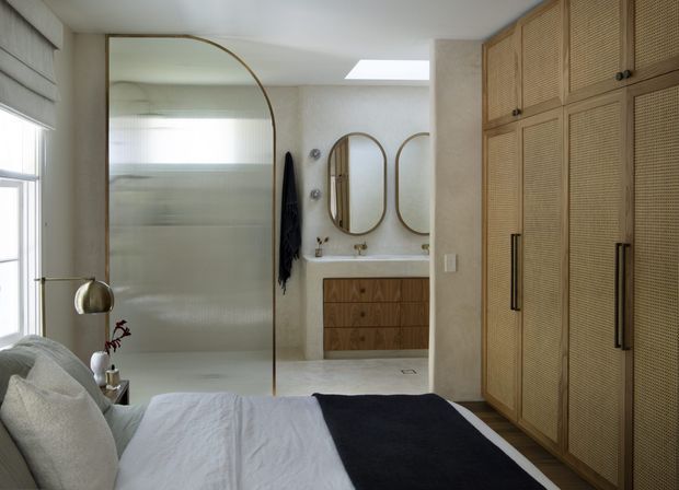 A refined bedroom with a partially open ensuite bathroom visible beyond a curved glass partition. The room features a neatly made bed, full-height woven-texture built-in wardrobes, a timber vanity with a white countertop, arched mirrors, pale textured walls, and a skylight. The palette is soft and neutral with dark blue-black accents.