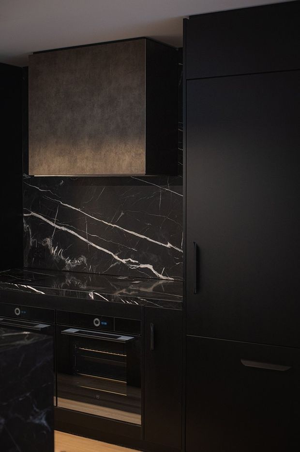 A dark, dramatic contemporary kitchen featuring floor-to-ceiling matte black cabinetry, integrated appliances, and a black marble or stone benchtop and splashback with prominent white veining. A warm textured bronze or taupe feature panel appears above the cooking area, while warm timber flooring adds contrast.