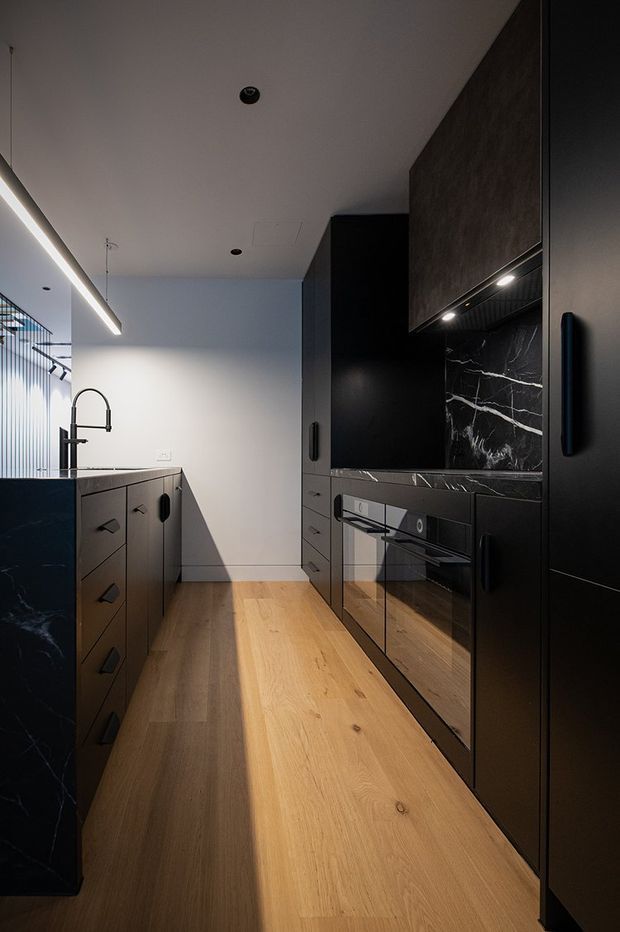 A narrow contemporary kitchen with matte black cabinetry, integrated appliances, dark veined stone splashback and benchtops, a black stone island, and warm natural timber flooring. A gooseneck sink tap and linear pendant light are visible, with strong contrast between the dark joinery and pale walls.