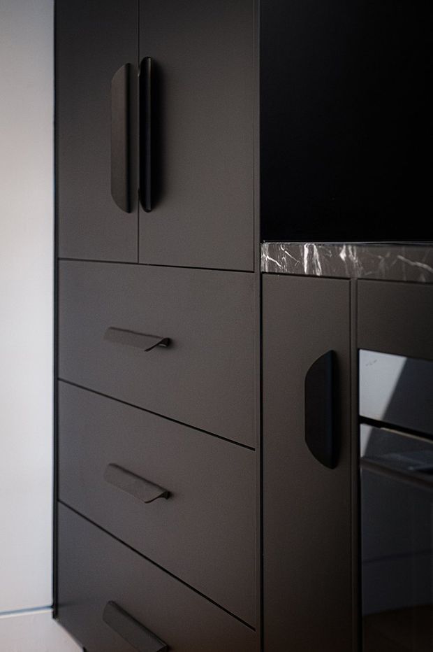 Close-up of dark matte built-in cabinetry with integrated drawers, tall cupboard doors, sculptural dark handles, and a built-in oven. A narrow veined stone or marble benchtop edge is visible, creating a refined, moody kitchen joinery detail.