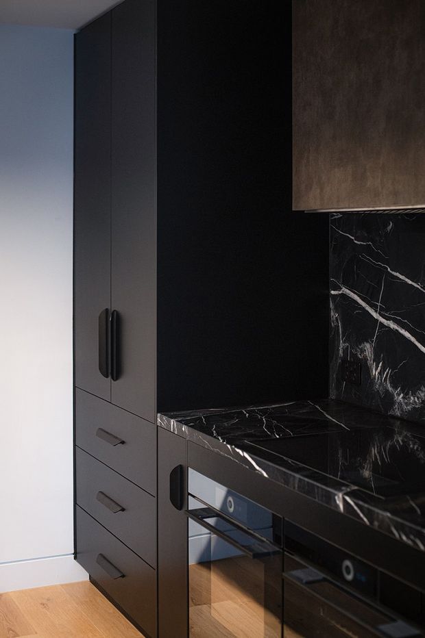A compact, dark-toned kitchen or kitchenette with full-height matte charcoal cabinetry, integrated drawers and appliances, and black marble-look benchtops and splashback with prominent white veining. Warm timber flooring contrasts with the black joinery, while a bronze-toned upper cabinet or rangehood surround adds a subtle metallic accent.