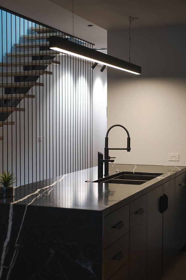 A moody contemporary kitchen with a large dark stone island, integrated double sink, tall black tap, dark cabinetry and a slim linear pendant light. Behind the kitchen is an open-riser staircase with timber treads and a black metal balustrade, creating a strong architectural feature.