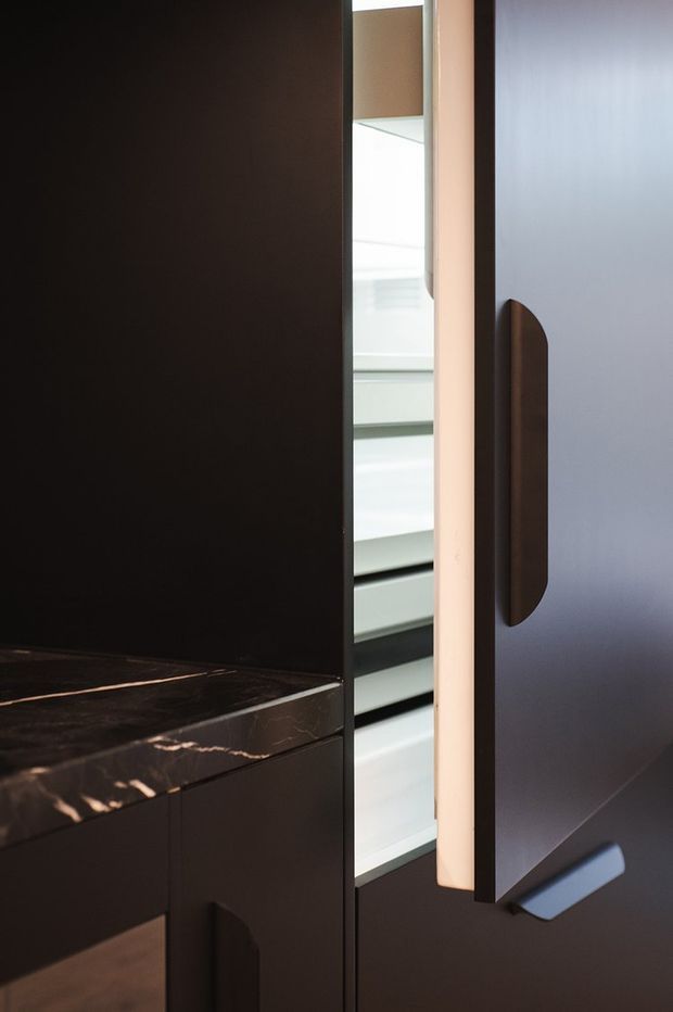Close-up detail of dark built-in cabinetry with a polished dark stone benchtop and integrated vertical handles. A tall cabinet door is partially open, revealing illuminated adjustable shelving inside. The composition is minimalist and high-contrast, focused on refined joinery and concealed storage.