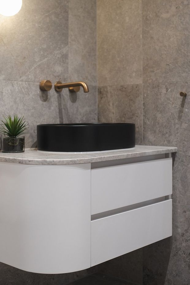 Close-up of a refined contemporary bathroom vanity. A matte black vessel basin sits on a light grey stone benchtop above a white floating drawer vanity with a curved left end. Grey stone-look wall tiles form the backdrop, with a brushed brass wall-mounted tap and a small potted plant adding detail.