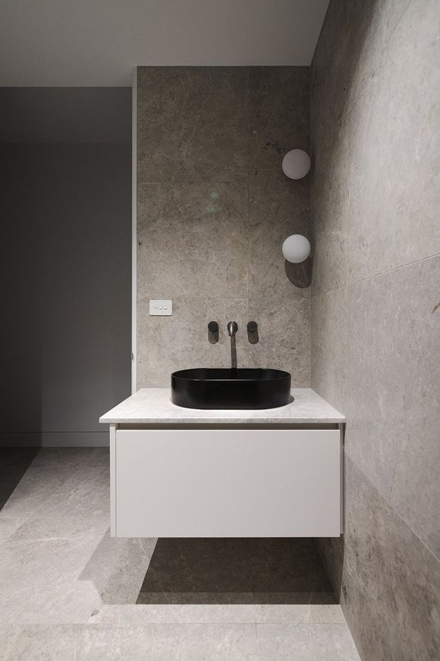 A compact contemporary bathroom featuring a wall-mounted white vanity with a pale stone benchtop, black vessel basin and black wall-mounted tapware. Large grey-beige stone-look tiles cover the walls and floor, with two spherical wall lights and a dark shower or doorway area visible to the left.