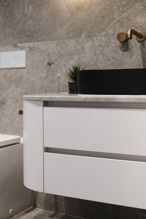 Close-up of a contemporary bathroom vanity with curved white drawers, a pale stone countertop, and a matte black vessel basin. The walls and floor are clad in grey stone-look surfaces, with a bronze wall-mounted tap and a small plant adding contrast.