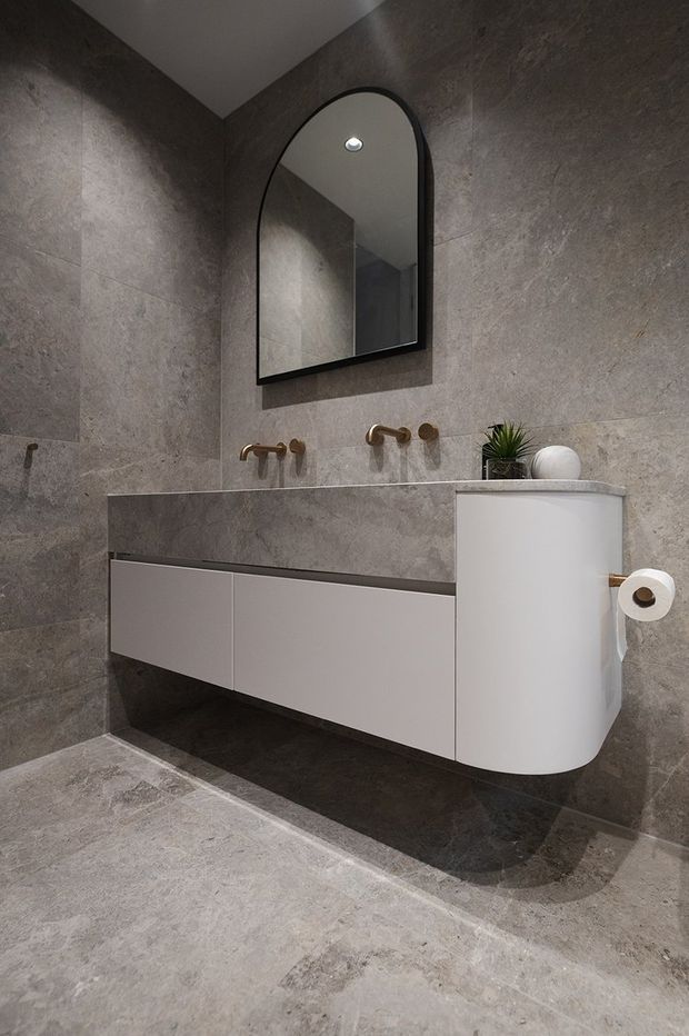 A refined contemporary bathroom featuring large-format grey stone-look wall and floor tiles, a floating white vanity with a curved end, a matching stone benchtop, brushed brass wall-mounted tapware, and a black-framed arched mirror. A small plant adds a subtle natural accent.