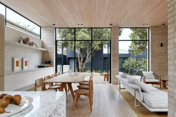A bright open-plan living and dining space with a large marble island in the foreground, a timber dining table and chairs, and a pale sectional sofa. Full-height black-framed glazing opens views to a landscaped courtyard, while a linear timber ceiling, pale timber flooring, textured light stone walls, and integrated white cabinetry create a warm contemporary interior.