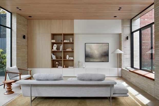 A bright, refined living room with a large light-grey sectional sofa facing a built-in media wall and open timber shelving. The room features pale timber cabinetry, exposed light brick walls, a warm timber slatted ceiling, large black-framed windows, and a neutral abstract artwork. A woven lounge chair and sculptural side table add texture, while abundant glazing creates an airy connection to the outdoors.