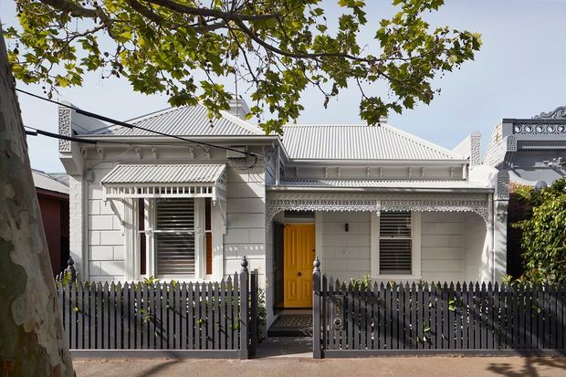 Street-facing view of a compact heritage-style single-storey house with pale horizontal weatherboard cladding, a corrugated pitched roof, ornate decorative trim, timber-framed windows with external blinds, and a bright yellow front door. A dark vertical picket fence and mature street tree frame the facade.