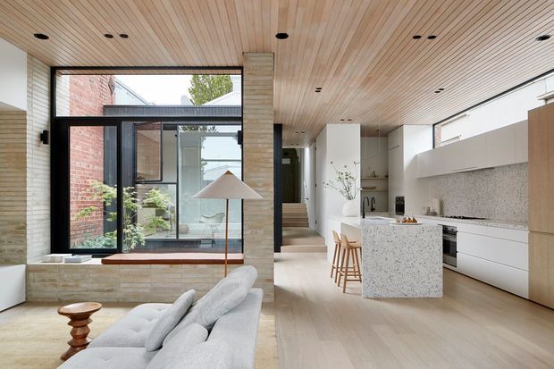 A bright, refined open-plan interior combining a contemporary kitchen, dining area and living space around a glazed courtyard. Pale timber slatted ceilings, light timber flooring, white cabinetry, a terrazzo island and backsplash, exposed brick, and dark-framed glazing create a warm minimalist atmosphere.