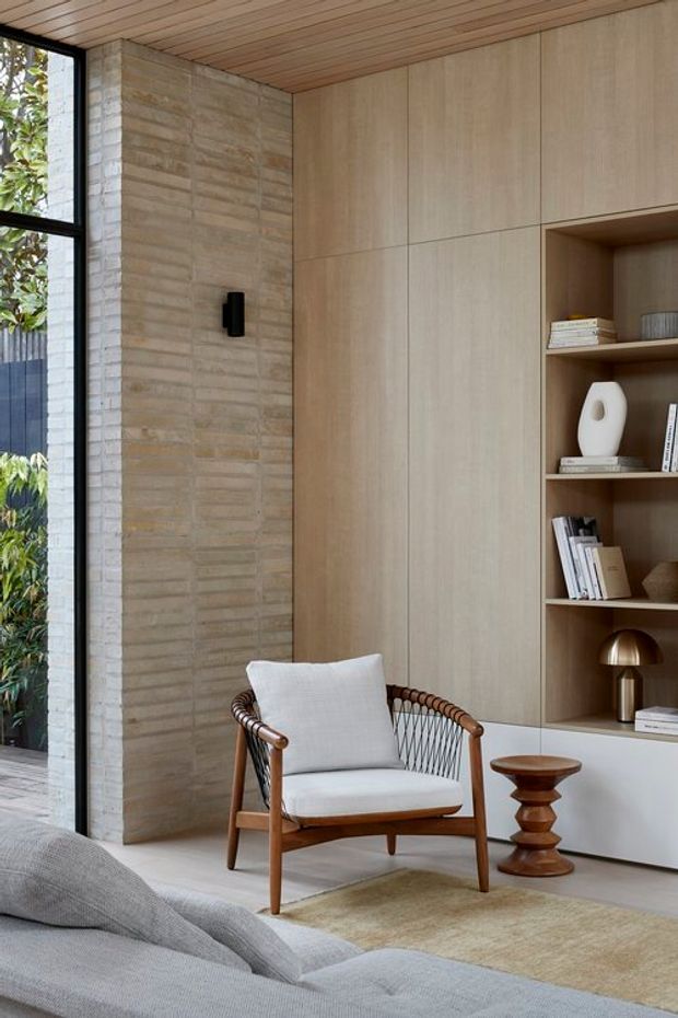 A refined living room interior with a mid-century-inspired timber and woven lounge chair, a large grey sofa in the foreground, pale timber built-in cabinetry and open shelving, a textured light brick feature wall, and a timber-lined ceiling. Floor-to-ceiling black-framed glazing brings in greenery from outside. Neutral natural materials and sculptural decor create a calm, contemporary setting.