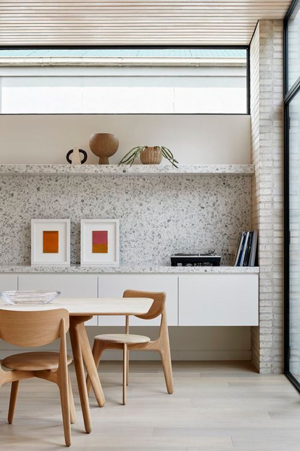 A bright, refined dining area with a light timber dining table and sculptural timber chairs, positioned in front of a built-in white sideboard. The sideboard features a large terrazzo-clad wall and benchtop with open display shelving, framed artwork, ceramics, books, and plants. Full-height glazing at the right, a high slatted timber ceiling, pale timber flooring, and a pale brick wall create a warm contemporary interior.