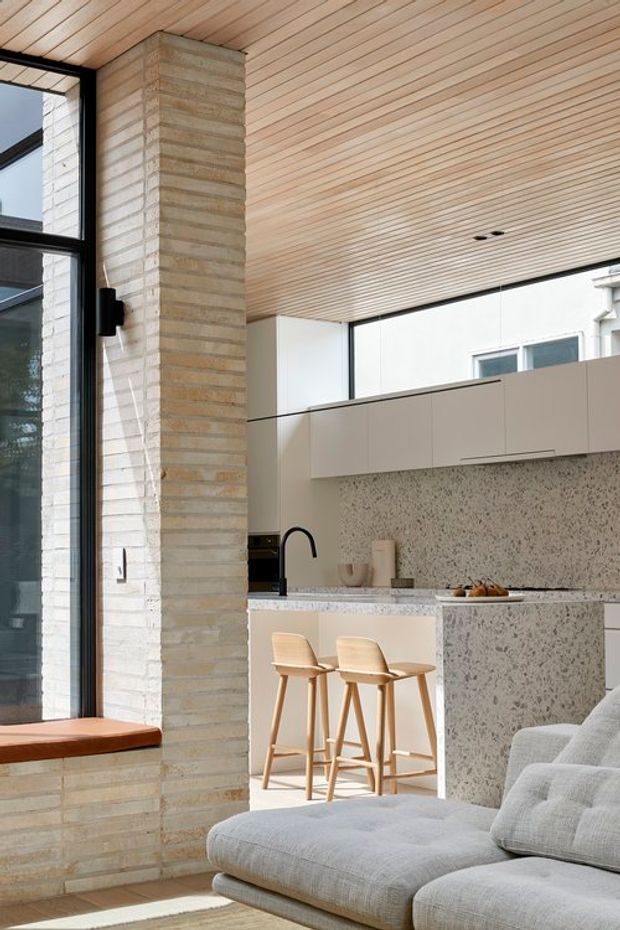 A bright open-plan kitchen and living area with a pale timber-lined ceiling, large black-framed glazing, and a prominent textured light brick column. The kitchen features light cabinetry, a speckled stone island and splashback, a black gooseneck tap, and two timber bar stools. A large light-grey sectional sofa occupies the foreground, creating a warm, refined contemporary interior.