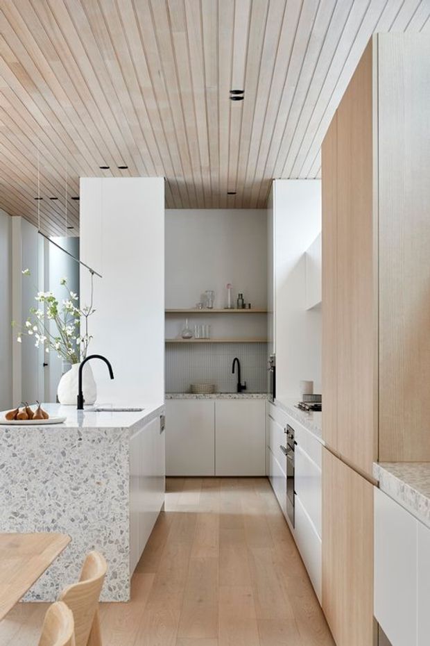 A bright, refined contemporary kitchen viewed down a narrow galley layout. The space features white cabinetry, light timber tall joinery, a pale terrazzo island and benchtops, integrated appliances, open shelving, black gooseneck and wall-mounted taps, and a linear light timber slatted ceiling. Floor-to-ceiling glazing at the side brings in abundant natural light, while a dining table and curved timber chairs are partially visible in the foreground.