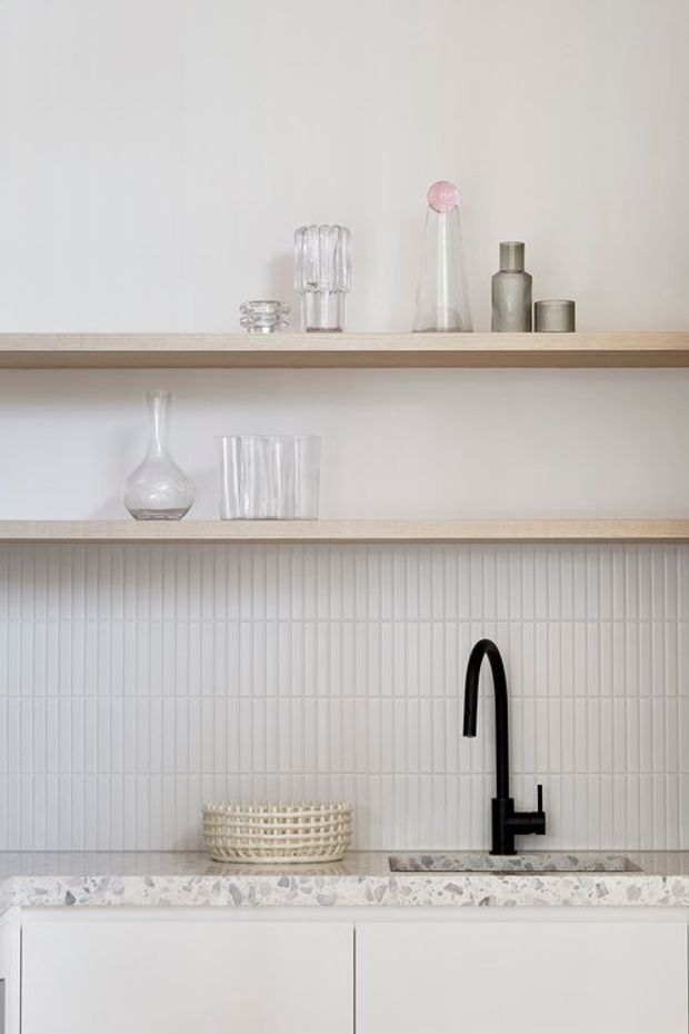 A refined minimalist kitchen vignette with white vertically fluted tile backsplash, pale timber open shelving, white cabinetry, a terrazzo-style benchtop, and a matte black gooseneck tap. Clear and muted glass vessels decorate the shelves, creating a calm, light-toned composition.