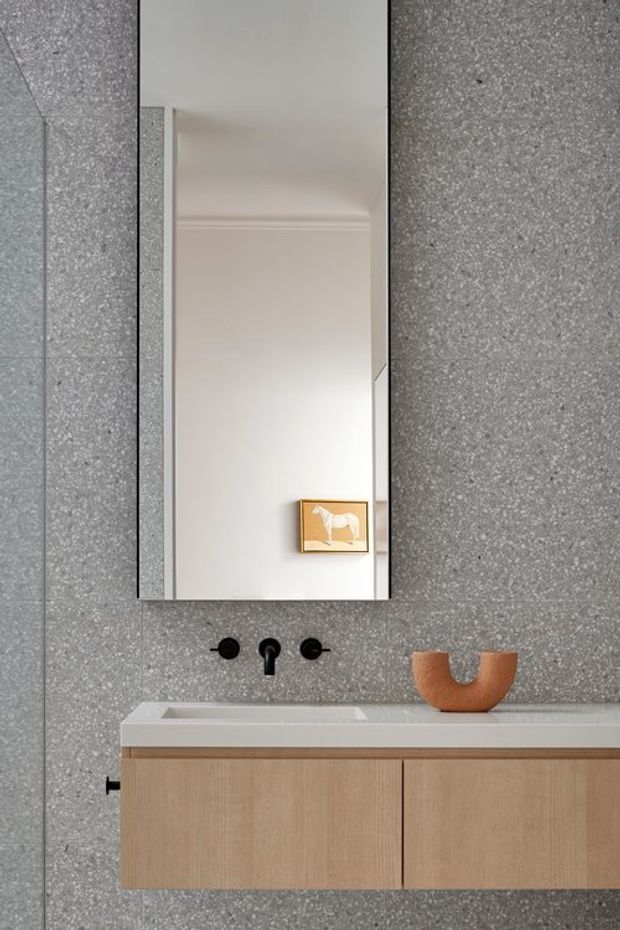 Minimal contemporary bathroom vanity with a floating light-wood cabinet, white integrated basin and benchtop, matte black wall-mounted tapware, and a tall frameless mirror. The walls are finished in speckled grey tile, with a small framed artwork and sculptural terracotta vessel adding restrained warmth.