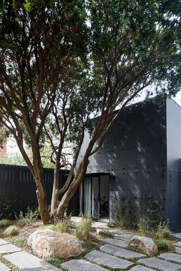 A contemporary dark-clad building partially screened by mature multi-trunk trees. A landscaped front garden features large natural boulders, ornamental grasses, gravel, and irregular concrete stepping stones leading to a glazed entrance. Vertical dark fencing and corrugated metal wall cladding create a restrained, modern exterior.