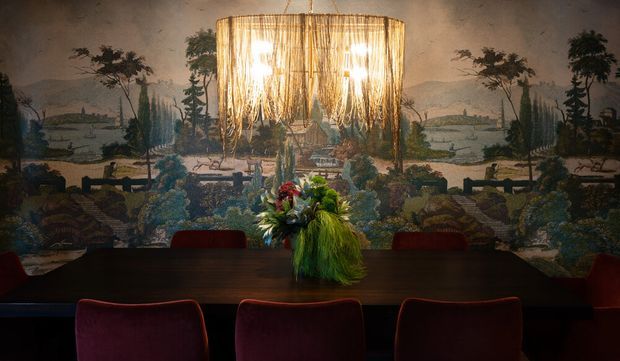 A moody formal dining room centered on a long dark dining table with deep red upholstered chairs, a dramatic oversized woven pendant chandelier, and a large scenic landscape mural covering the rear wall. A vivid floral arrangement forms the table centerpiece.