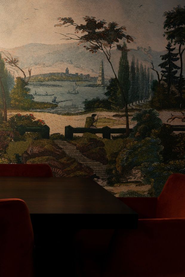 A dimly lit interior dining area with a dark rectangular dining table and deep red upholstered chairs in the foreground. The wall behind the table is covered by a large scenic mural or printed wallpaper depicting a tranquil lake, sailboats, trees, landscaped grounds, and a distant tower or village. The space has a dramatic, moody atmosphere with rich dark tones and a historic, pictorial character.
