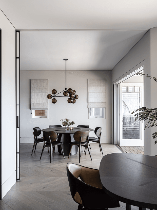 Contemporary dining area with a round dark dining table and sculptural black-and-brown dining chairs, set against light grey walls and herringbone timber flooring. A clustered globe pendant hangs above the table, with two small windows fitted with woven Roman blinds. Full-height glazed doors open to an exterior brick facade, while a second dark table and chair are visible in the foreground.