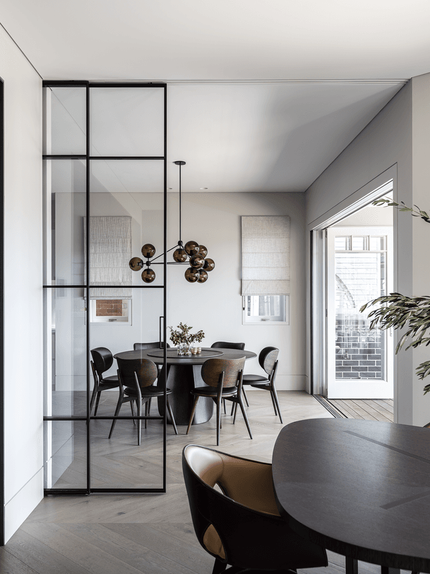 A refined contemporary dining area with a round dark dining table and matching chairs, viewed through a black-framed glass partition. The room features pale timber flooring, light neutral walls, roller blinds, a sculptural clustered pendant light, and full-height glazed doors opening to the exterior. A second table and chair are visible in the foreground, with a leafy indoor plant at right.