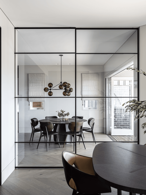 A refined dining room viewed through a black-framed, full-height glass partition. The room features a round dark dining table with sculptural dark chairs, a contemporary globe pendant, pale walls, textured window coverings, and light herringbone timber flooring. A bright entry or exterior doorway is visible beyond the dining area, while a portion of an adjacent dark table and chair appears in the foreground.
