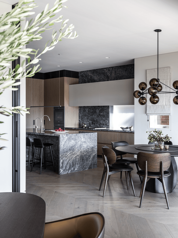 A refined open-plan kitchen and dining area with dark timber cabinetry, a dramatic veined stone island and backsplash, integrated appliances, and a round dining table with curved upholstered chairs. A sculptural clustered pendant light hangs above the dining area, while a large leafy branch frames the foreground. The space has a sophisticated, contemporary feel with herringbone timber flooring and neutral finishes.