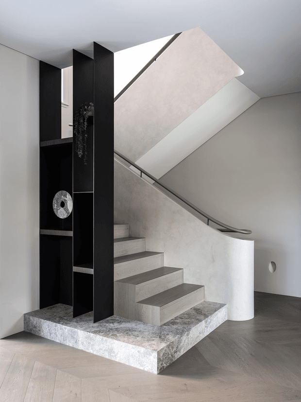 A sculptural contemporary stairway with pale stone or concrete steps rising around a curved white balustrade. A dark, floor-to-ceiling built-in shelving partition frames the stairs, while a large angled ceiling opening and soft plastered surfaces create a dramatic architectural composition. The floor appears to be light timber in a herringbone pattern, with restrained monochromatic styling.