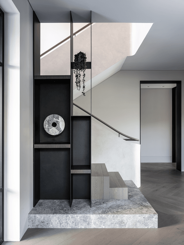 Contemporary interior entry and stair hall featuring a sculptural staircase, dark built-in display shelving, a stone plinth, expansive mirror panels, black-framed openings, and a restrained monochromatic palette. A hanging plant and decorative object add subtle organic detail.