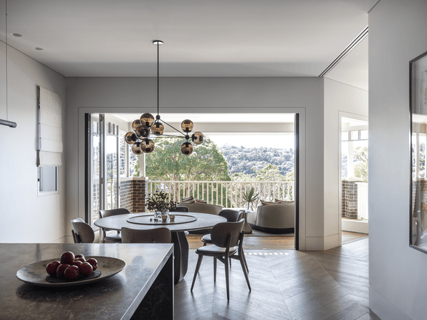 A refined open-plan dining and living area with a dark stone kitchen island in the foreground, round dining table and chairs beneath a sculptural globe pendant, and a lounge seating area beyond. Large glazed openings lead to a balcony with a balustrade and leafy views. The interior uses neutral grey walls, herringbone timber flooring, dark cabinetry or stone surfaces, and contemporary mid-century-inspired furnishings.