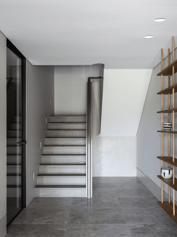 A contemporary stairway and landing with grey stone-look tiled flooring, grey walls, pale timber stair treads, and a sleek curved metal-and-glass balustrade. A full-height dark-framed glazed opening is visible to the left, while open timber shelving with decorative ceramics lines the right wall. Recessed ceiling lights create a clean, minimalist feel.