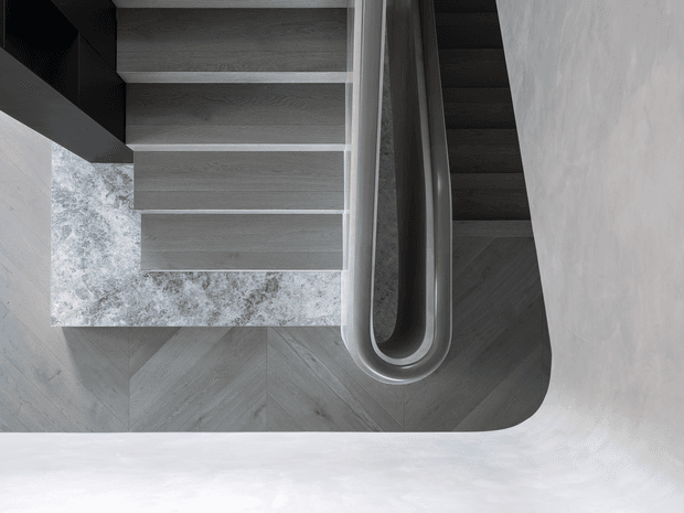 Top-down view of a contemporary staircase and landing, featuring dark grey timber-look treads, a sculptural rounded stair or balustrade edge, and grey chevron-patterned flooring. The monochromatic composition is softened by light textured walls and a mottled grey feature surface.