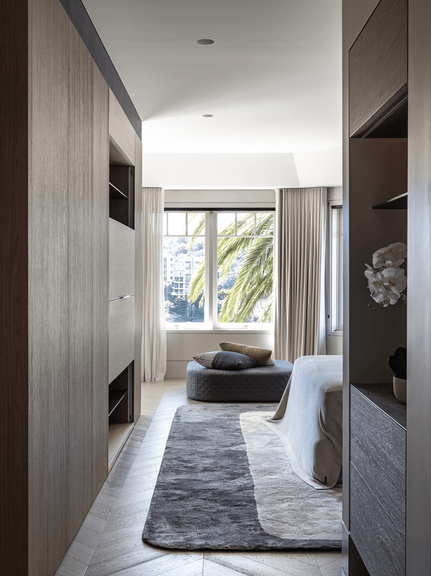 A refined contemporary bedroom viewed through a narrow passage between full-height timber-toned built-in joinery. The room features a partially visible upholstered bed, a large two-tone area rug, a low tufted ottoman, floor-to-ceiling curtains, and a bright window overlooking palm foliage and nearby buildings. The neutral palette and concealed storage create a calm, sophisticated interior.