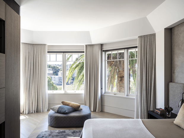 Bright contemporary bedroom with a large upholstered bed, tufted ottoman, full-height pleated curtains, and expansive corner windows overlooking palm trees and a residential landscape. The room features pale wood wall cabinetry, white walls and ceiling, light timber flooring, and restrained neutral furnishings.