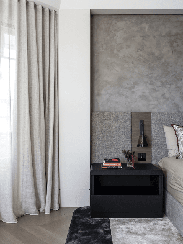 A refined contemporary bedroom corner featuring a large window with full-height textured beige curtains, pale timber flooring, and a dark black bedside table. The bed is partially visible at right with neutral bedding and cushions. Behind the bedside table is a layered feature wall combining grey textured plaster, grey upholstered wall panels, and a vertical dark timber panel with integrated lighting and controls. A two-tone black and light-grey rug adds contrast.