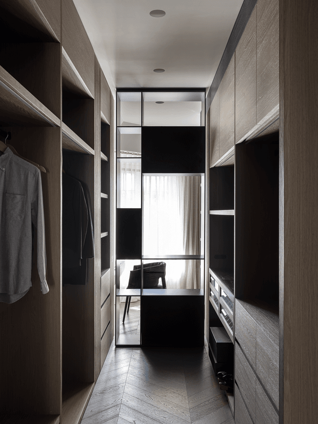 A narrow, refined walk-in wardrobe with full-height dark timber cabinetry and open shelving on both sides. Clothing hangs in the left alcoves, while drawers and storage compartments line the right side. A black-framed glazed partition at the end reveals a bright seating area with sheer curtains and a chair. The floor features a chevron timber pattern, and recessed ceiling lights provide subtle illumination.