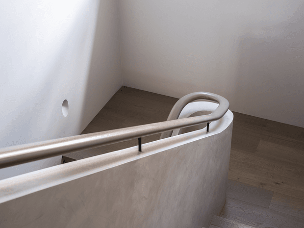 Minimalist interior view of a curved stairway or landing with a smooth plastered balustrade, rounded metal handrail, light wood flooring, and plain white walls. The composition emphasizes the sculptural curve of the staircase and restrained material palette.